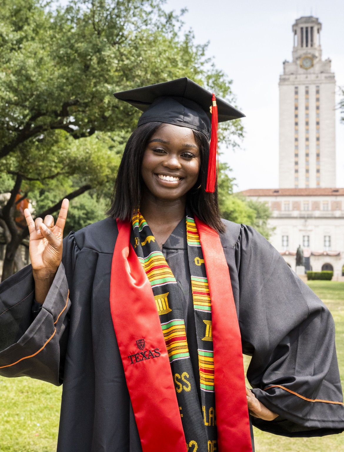 Student in graduation regalia in front of the UT tower, showing hook 'em hand signal