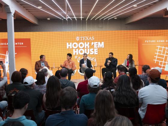 group of people on a panel in front of a crowd in chairswith a burnt orange hook 'em house logo in the background on a screen