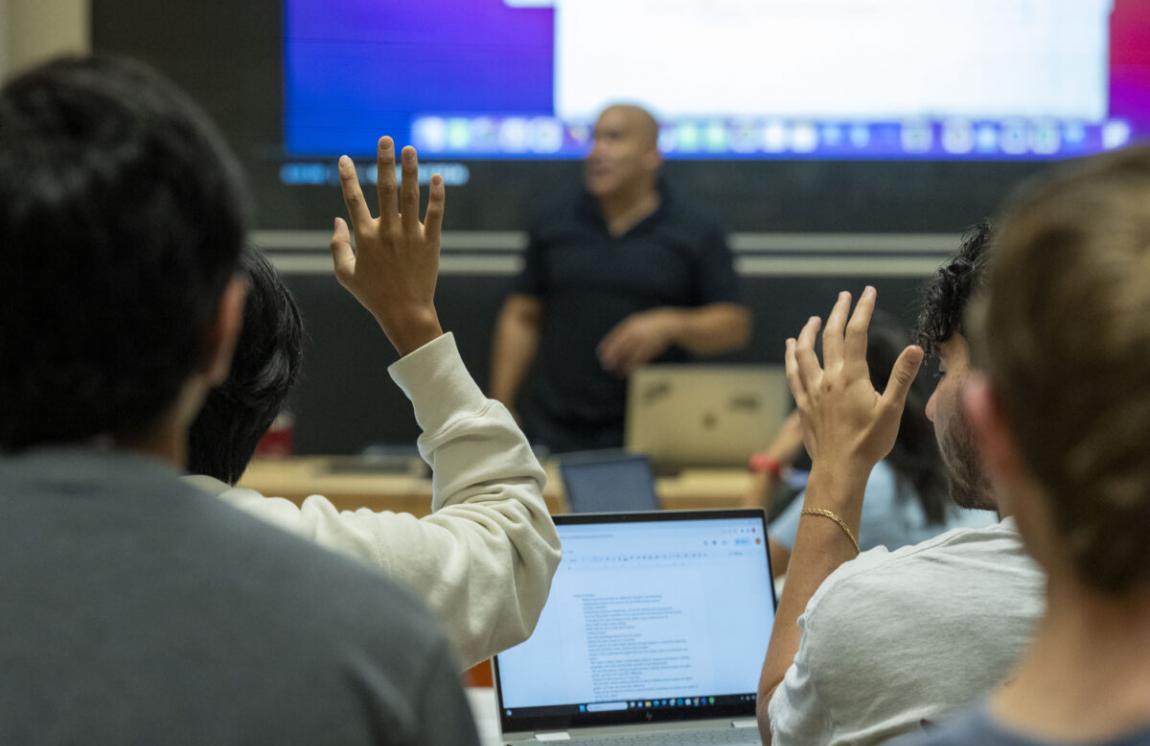 students in a classroom with hands raised and professor teaching in background