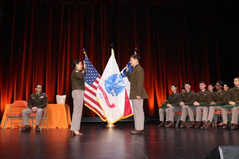 Two military officers in uniform conducting a formal flag ceremony on stage, with additional uniformed personnel seated in the background and U.S. and military flags displayed.