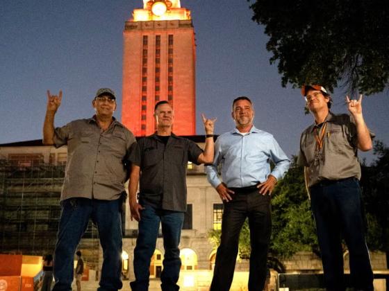 Four men stand in front of the University of Texas at Austin Tower at night as it glows orange.