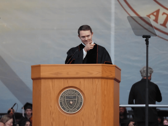 Former UT quarterback Colt McCoy points to the crowd from a podium while wearing a black graduation robe.