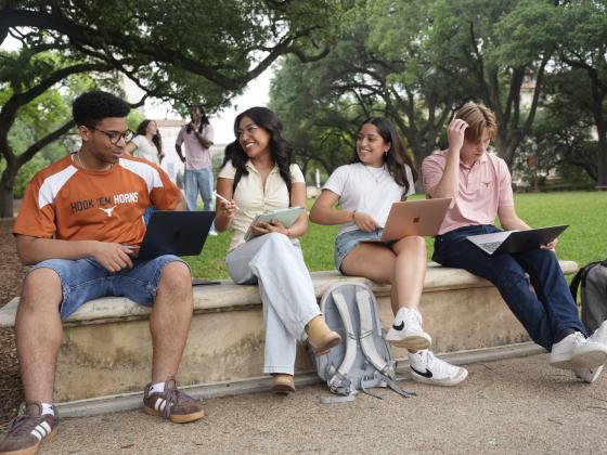 Students with laptops and tablets smile while talking on a bench on the University of Texas at Austin campus.