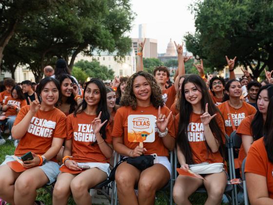 A group of students wearing burnt orange T-shirts smile and hold up the Hook 'em, Horns hand sign while posing for a photo.