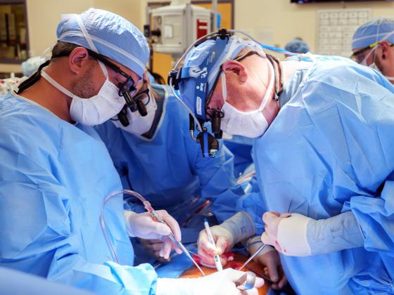 Three surgeons, all wearing blue scrubs and protective equipment, doing a surgical procedure in an operating room.