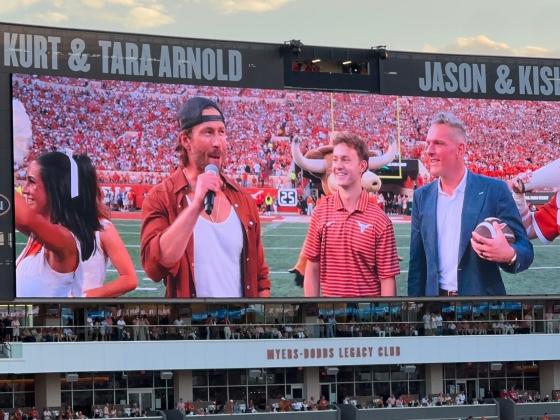 On a stadium screen, a UT student and College GameDay analyst Pat McAfee look on as actor Glen Powell speaks into a microphone.