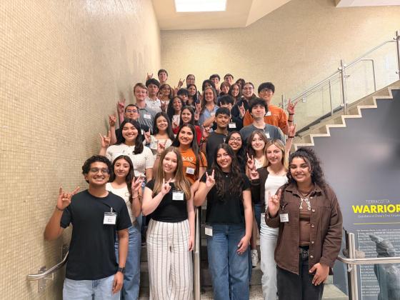 A group of students standing on a staircase smile and hold up the Hook 'em, Horns hand sign while posing for a photo.