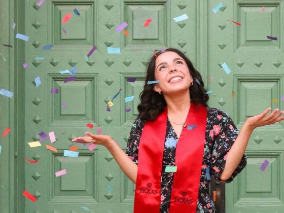 A female student wearing a red graduation stole smiles in front of green double doors as confetti falls around her.