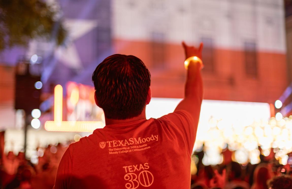 A photo of the back of a student at an event wearing a burnt orange T-shirt and holding up the Hook 'em, Horns hand sign.