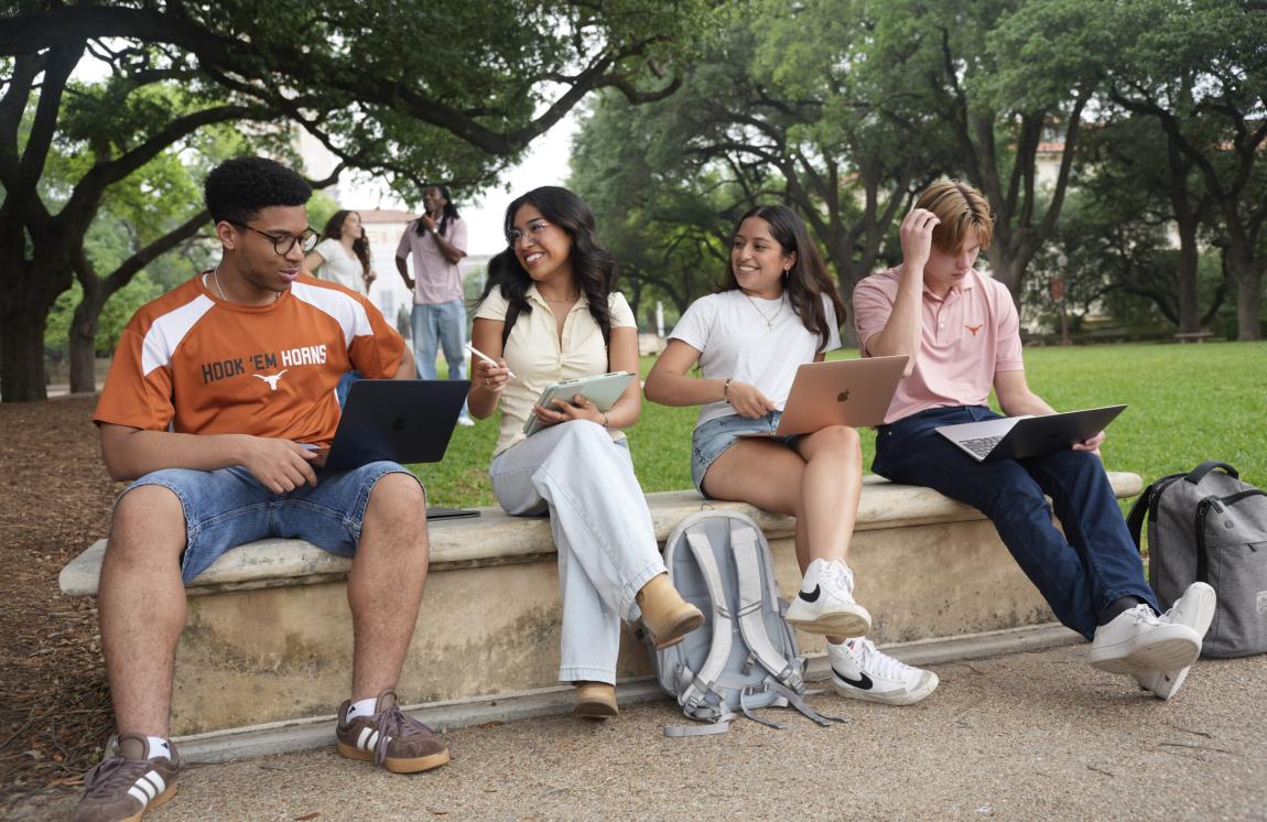 Students with laptops and tablets smile while talking on a bench on the University of Texas at Austin campus.