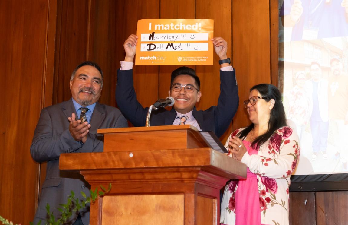 A medical student stands at a podium with his family while holding a sign that says "I matched in Neurology at Dell Med."