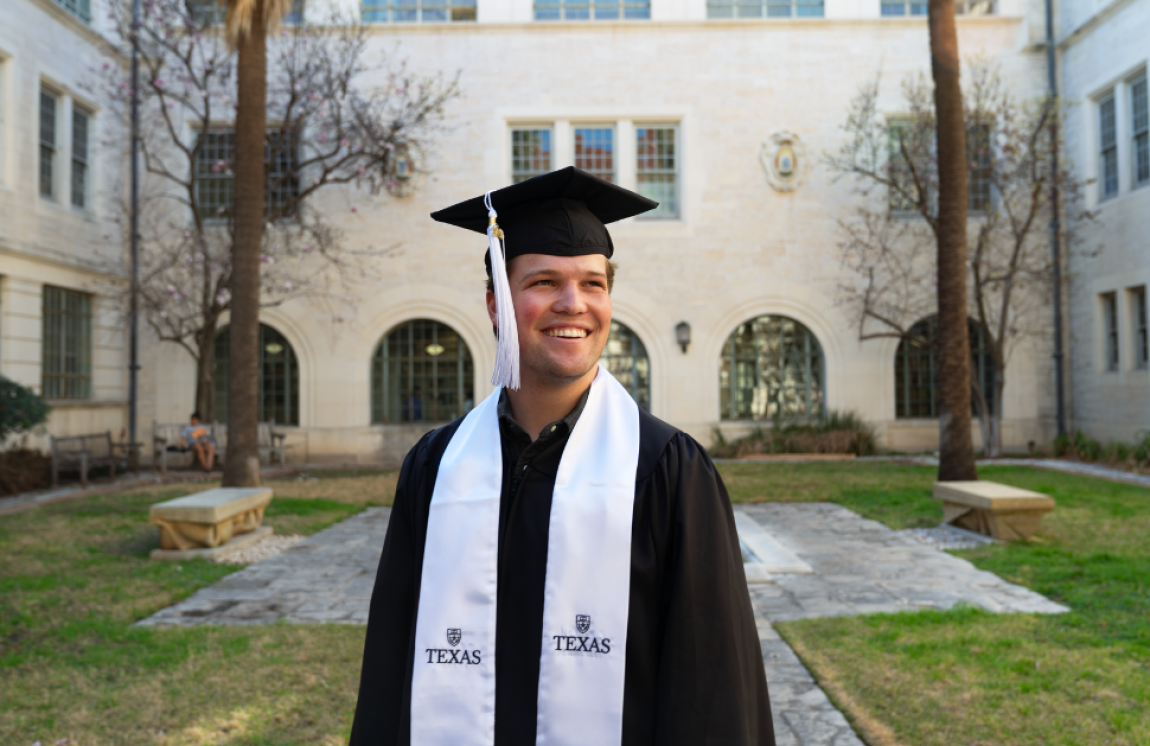 A UT graduate smiles while wearing a black graduation cap with a white tassel and a black graduation gown with a white stole.