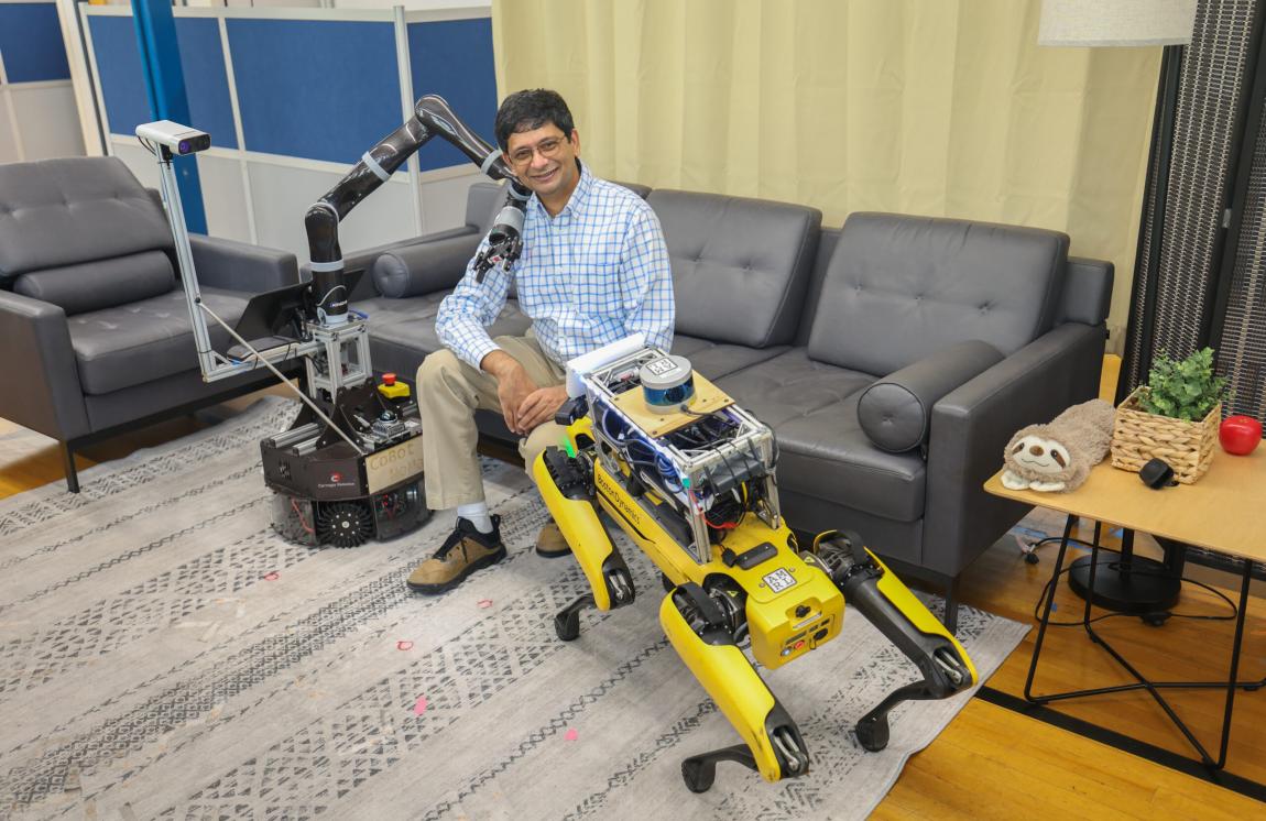 A man smiles for a photo while sitting on a couch with robots on either side of him.