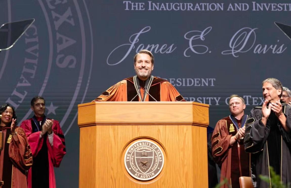 President Jim Davis at podium in regalia smiling with other people standing on stage in regalia applauding