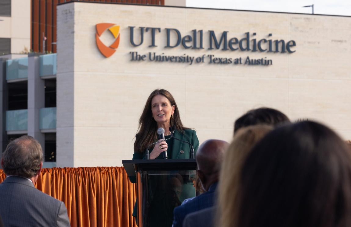 A woman speaks into a microphone while addressing an audience. Behind her is a building with the UT Dell Medicine logo.