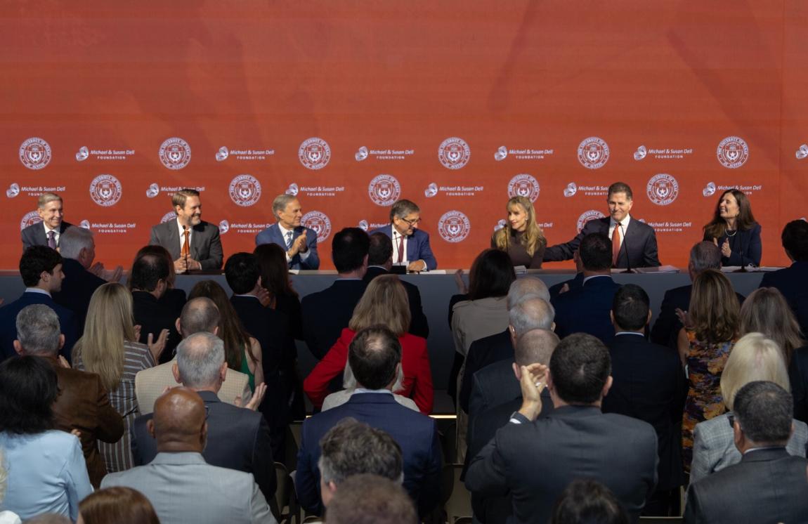 A panel of seven people sits at a long table on a stage with a burnt orange backdrop with logos.