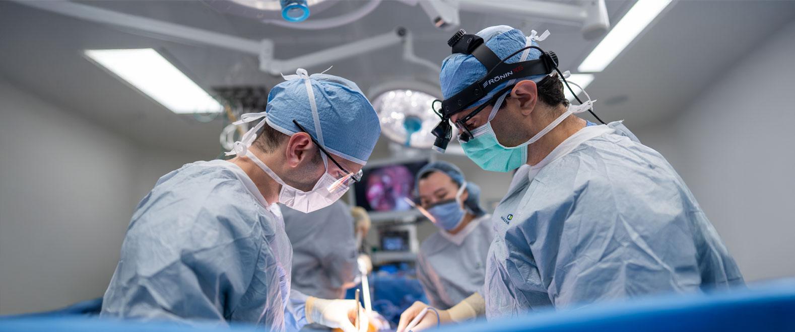 Surgeons in blue surgical scrubs and masks perform a procedure in a modern operating room, working under bright lights with a medical team in the background.
