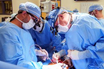 Three surgeons, all wearing blue scrubs and protective equipment, huddle close together during a surgical procedure in an operating room.