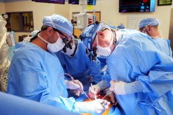 A surgical team in blue gowns, masks, and head coverings leans over a patient, performing a procedure under bright operating-room lights.