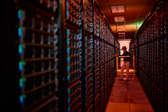 A technician works inside a data center aisle lined with tall server racks on both sides. Warm orange lighting contrasts with the cool blue indicator lights on the servers as the person adjusts equipment, highlighting the scale and intensity of modern computing infrastructure.