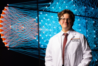 A smiling person wearing glasses and a white lab coat stands in front of a large digital display featuring an intricate network of blue and orange interconnected nodes and lines. They are dressed in professional attire with a tie, and the high-tech background suggests research, data science, or advanced scientific analysis.