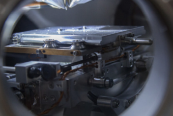 A close-up view of advanced laboratory equipment featuring a precision mechanical stage and mounted sample platform, seen through a circular frame. The setup includes metallic components, fine adjustment mechanisms, and wiring, suggesting materials science research or high-precision experimental analysis in a controlled lab environment.