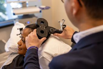 A man holds a medical instrument up to a woman's head. The woman is reclining in a patient bed.