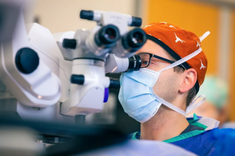 A medical professional closely observes through a surgical microscope in a clinical setting. The individual is wearing a burnt orange surgical cap with white Longhorns and a face mask, indicating a sterile environment. The background is softly blurred, focusing attention on the advanced optical equipment and the surgeon's concentration.
