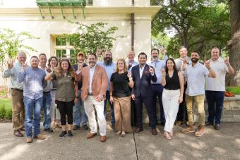 A group of roughly twenty people posing outdoors in front of a building with lush greenery, several giving the Hook 'em Horns sign, dressed in business casual attire.