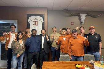 A group of people in a room decorated with a framed UT Austin jersey and Longhorns memorabilia, several giving the Hook 'em Horns sign, posing together for a photo.