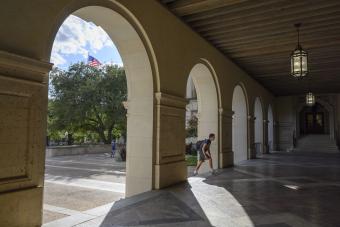 A view through a series of arched columns in a covered walkway, looking out onto a sunlit campus courtyard with trees, a flagpole flying an American flag, and hanging lantern-style light fixtures along the ceiling.