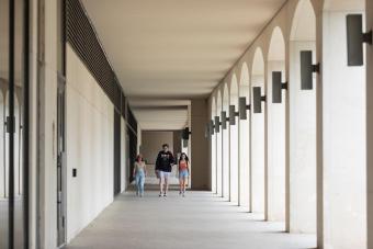 Three students walk together down a long, arched breezeway on a university campus, with a row of light fixtures mounted between columns leading into the distance. One wears a burnt-orange Texas cap and a Texas-branded sweatshirt, evoking a University of Texas at Austin campus setting.