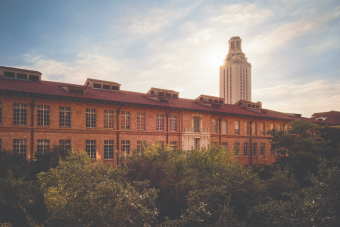 A red-brick, red-tile-roofed campus building with rows of tall windows and dormer vents, with the University of Texas at Austin Tower rising behind it, backlit by sunlight, against a partly cloudy blue sky, with dense green trees in the foreground."