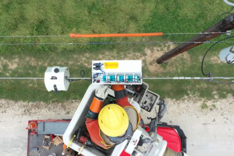 A view from above of a utility worker in a vest and hard hat attaching a blue sensor to a power line.