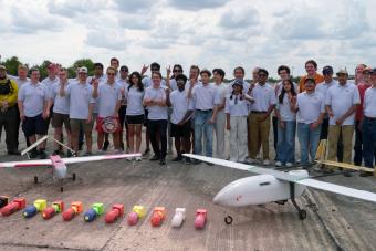 A large group of students and researchers pose on a concrete runway with two fixed-wing drones and a row of colorful test payloads on the ground in front of them. Most of the people in the photo are wearing matching light grey polo shirts, and several are making the "Hook 'em, Horns" hand gesture.