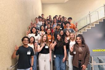 A group of students standing on a staircase smile and hold up the Hook 'em, Horns hand sign while posing for a photo.