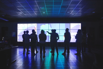 A group of people standing in front of a large, multi-screen data visualization display wall inside an interactive collaboration or research lab.