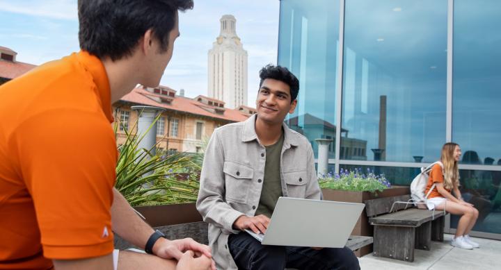 Two students talk on a balcony with the University of Texas at Austin Tower in the background.
