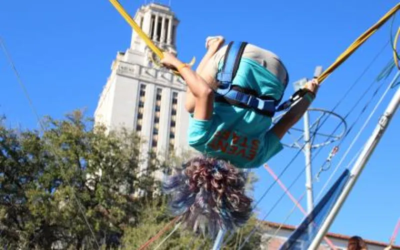 A student hangs upside down while attached to bungee ropes at a carnival attraction. Behind them is the UT tower.