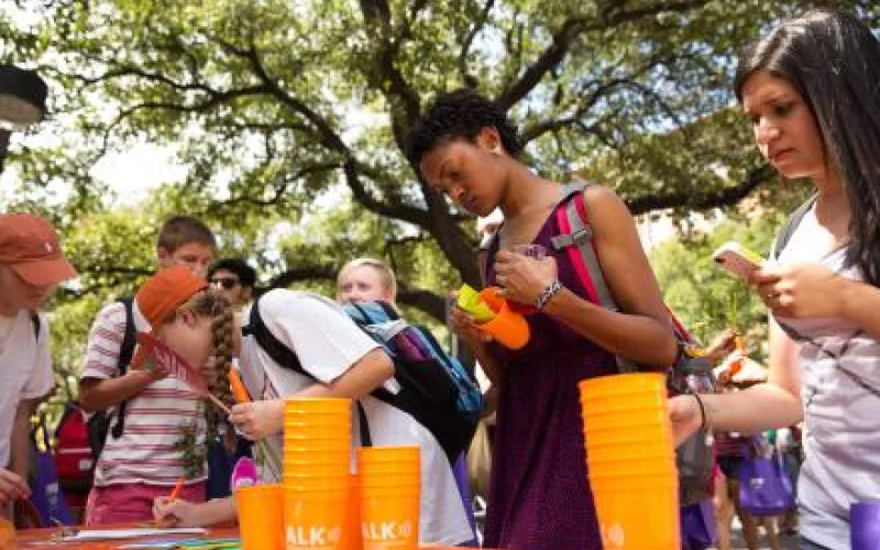 Several students gather at an organization tabling on campus with orange stacked cups. One girl writes on the table's paper and another holds one of the cups as she reads about the group.