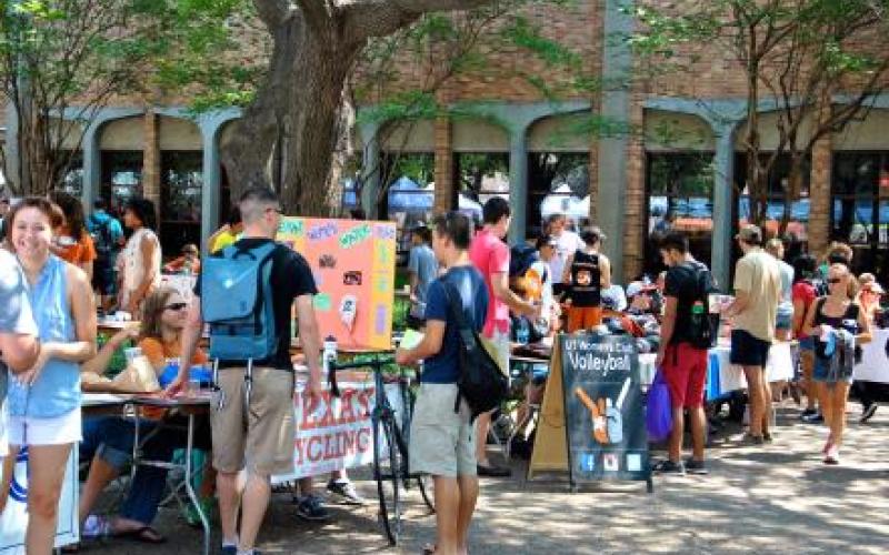 Many different tables for student organizations sit outside on the plaza. They're brightly colored with banners advertising their organizations, and many students walk between the tables and talk to the students behind them.