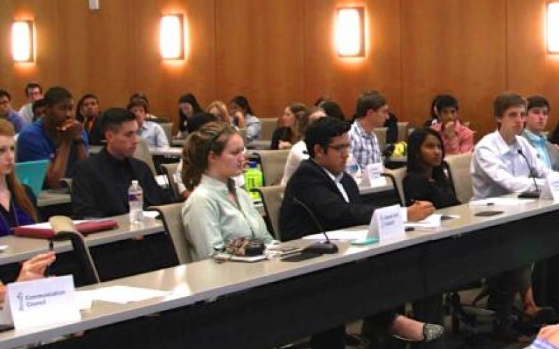 Around fifteen students dressed in professional clothes sit in four rows at a student government meeting. They look towards the front of the room.