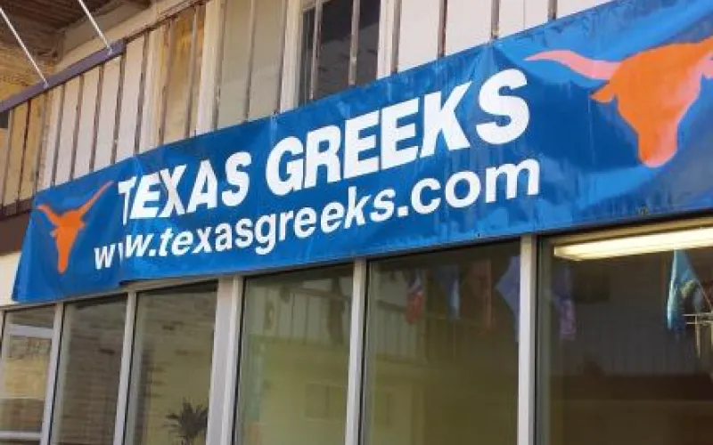 A big blue banner with the words Texas Greeks, a website URL, and two longhorns, hangs on the side of a beige building.