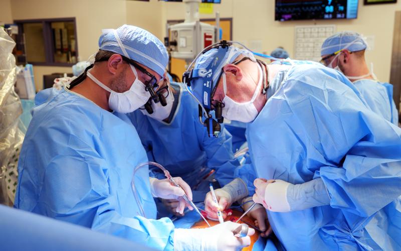A surgical team in blue gowns, masks, and head coverings leans over a patient, performing a procedure under bright operating-room lights.