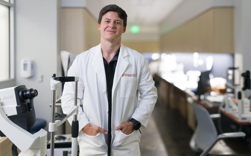 A clinician in a white lab coat stands with hands in pockets next to an eye exam machine in a clinic hallway, with workstations blurred in the background.