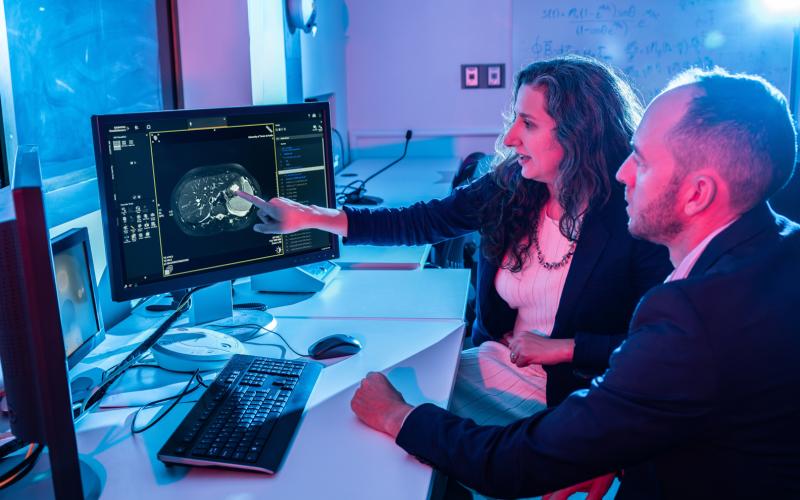 A researcher points to a cross-sectional abdominal scan on a large computer monitor while a colleague watches from beside the desk in a clinical lab.