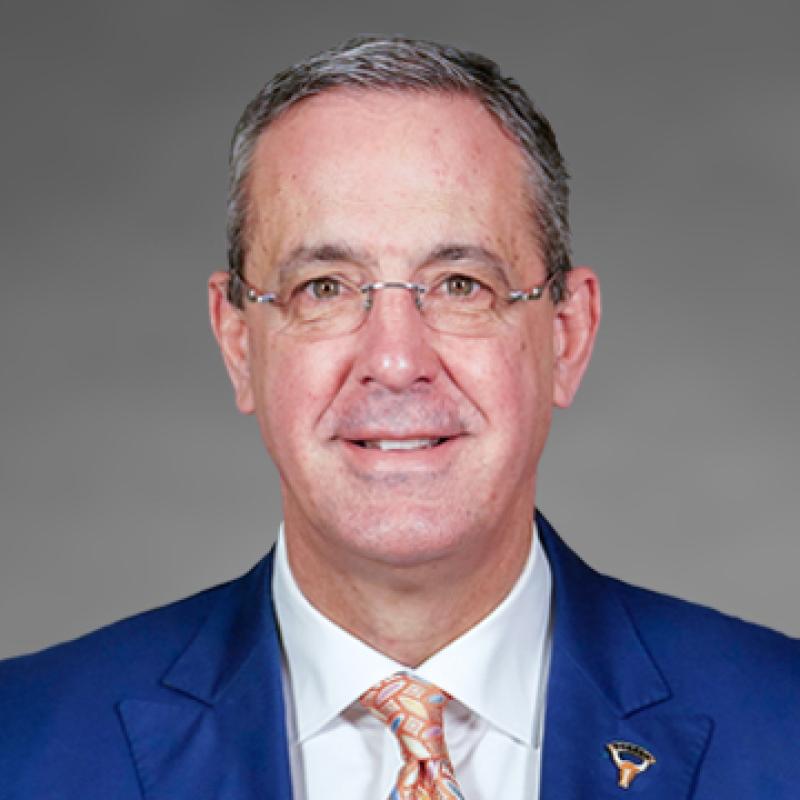 Headshot of Chris Del Conte, smiling, wearing glasses and a bright blue blazer with an orange tie, photographed against a gray background.