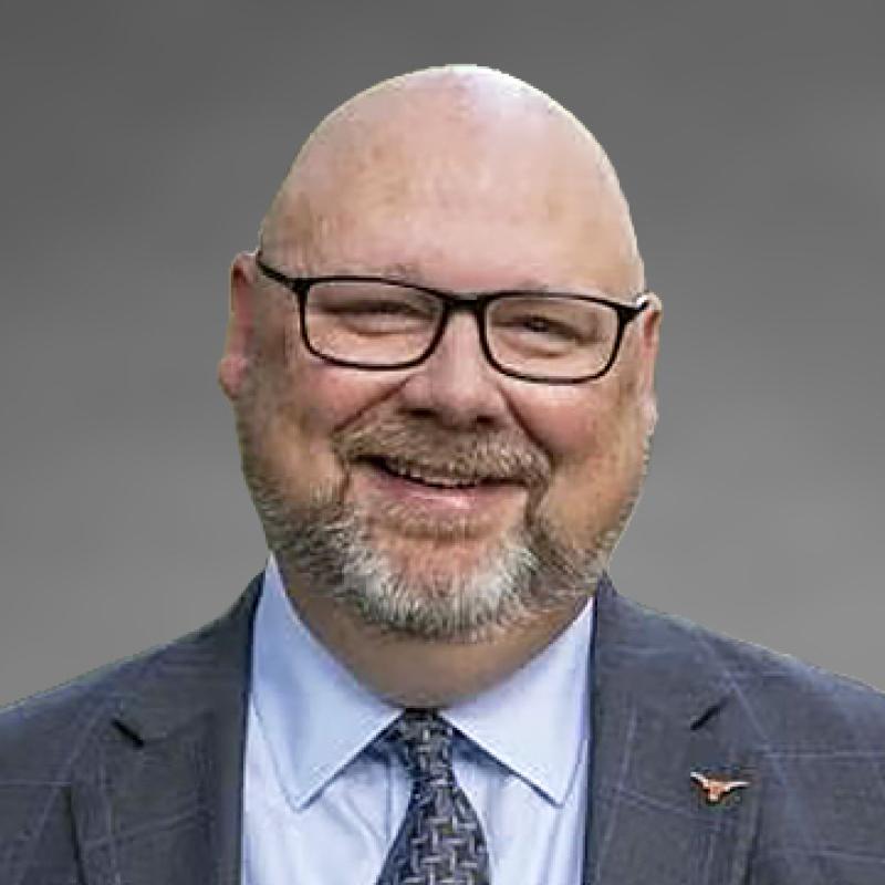 Headshot of Cole Camplese, smiling, with glasses and a beard, wearing a gray blazer, light blue shirt, and patterned tie with a small Longhorns pin on his lapel, photographed against a gray background.