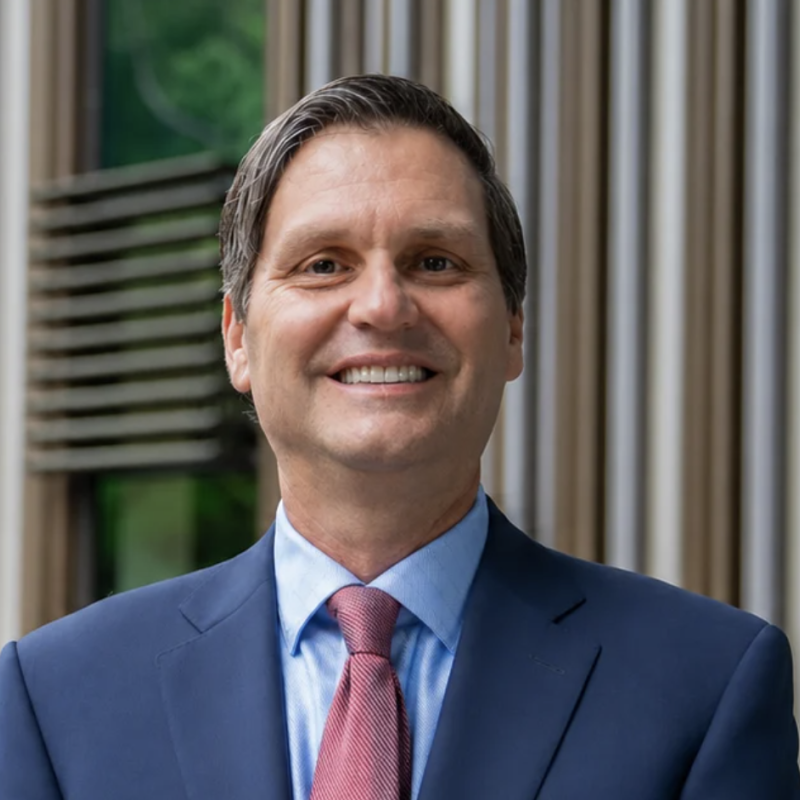 Bradley Staats in a navy blue suit, light blue dress shirt, and red tie, photographed outdoors in front of architectural columns with greenery in the background.