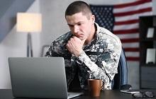 Photo of a male soldier in uniform studying at a laptop with an American flag in the background